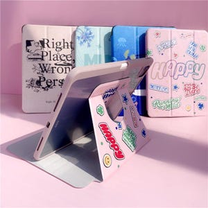 Könnte beinhalten: Eine rosa Tablethülle mit einem ausklappbaren Ständer. Die Hülle ist mit verschiedenen bunten Aufklebern mit Texten wie "Happy", "Another Level", "Running Wild" und "Heart on the Window" verziert.