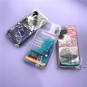 Puede incluir: Tres fundas transparentes para teléfono con diseños decorativos. Una funda presenta un diseño de "Magic Shop", otra tiene el texto "YOU NEVER WALK ALONE", y la tercera muestra un carrusel con el texto "Spring Day". Cada funda tiene un interior lleno de líquido y purpurina.