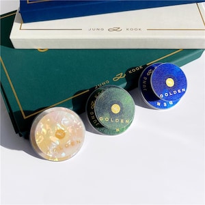 May include: Three round, iridescent, and blue-toned decorative items with the word "GOLDEN" printed on them. The items are on a green surface with a white box and the text "JUNG KOOK" visible.