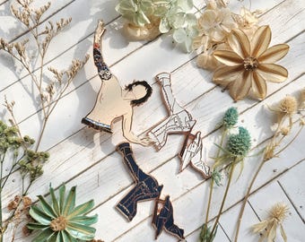 May include: Three gold-toned metal pins depicting a person in a variety of poses. The pins are on a white wooden surface with dried flowers and plants.