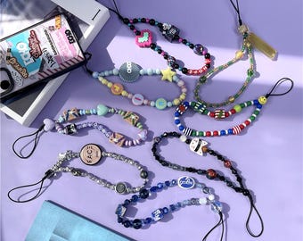 May include: A selection of beaded phone charms in diverse colours and designs. Some charms feature heart, star, and smiley face beads, while others have lettered beads spelling out words. The charms are on a purple surface.