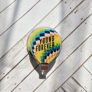 May include: A colorful hot air balloon-shaped object with the words "YOUNG FOREVER" printed on it. The balloon features a checkered pattern in shades of blue, green, yellow, and orange. The object is set against a white wooden background.