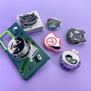 May include: A collection of phone grips and a phone case. The grips feature various designs, including a black and white image, a number seven, and a robot. The phone case is green with a yellow border and the text "SO COOL".