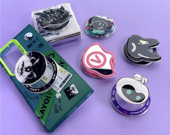 May include: A collection of phone grips and a phone case. The grips feature various designs, including a black and white image, a number seven, and a robot. The phone case is green with a yellow border and the text "SO COOL".