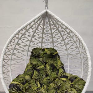 Puede incluir: Silla colgante de huevo de mimbre blanco con un cojín estampado de hojas tropicales negras y verdes.