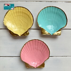 Op de afbeelding: Drie geschilderde schelpen in geel, turquoise en roze. Elke schelp heeft een goudkleurige rand en basis. De schelpen zijn gerangschikt op een wit houten oppervlak. In de linkerbovenhoek staat een bloemenontwerp met de tekst "NUMBER 45".