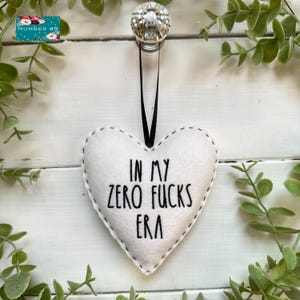 Può includere: Ornamento a forma di cuore in feltro bianco con bordi cuciti neri e la frase "IN MY ZERO FUCKS ERA" in lettere nere. L'ornamento è appeso a un nastro nero. La vegetazione decorativa circonda l'ornamento.