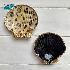 May include: Two decorative seashell-shaped dishes. One has a leopard print design with gold trim, the other is solid black with gold trim. They are on a white wooden surface. The text "NUMBER 45" is in the upper left corner.