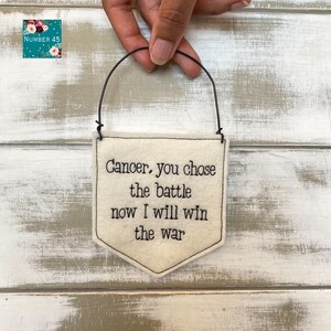 以下が含まれることがあります： 黒い刺繍で「癌、あなたは戦いを決めた、今私は戦争に勝つ」と書かれた白いフェルトの旗。旗は黒いワイヤーのループから吊り下げられています。