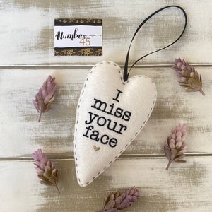 May include: White felt heart-shaped ornament with black embroidery that reads "I miss your face". The ornament has a black ribbon loop for hanging and a small gold glitter heart in the centre.