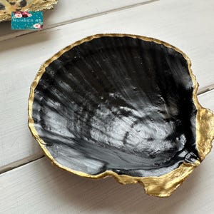 May include: A decorative, shell-shaped dish with a black interior and a gold-painted rim. The dish has a textured surface and a unique, organic shape. The dish is likely used for holding small items or as a decorative accent.