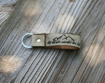 May include: A tan leather keychain with a silver key ring. The keychain has the phrase "Not all who wander are lost" and a mountain graphic etched into the leather. The keychain is on a wooden surface.