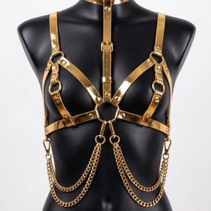Puede incluir: Arn&eacute;s corporal dorado con un collar tipo gargantilla. El arn&eacute;s presenta correas ajustables, anillos met&aacute;licos y cadenas decorativas. El dise&ntilde;o se muestra en un maniqu&iacute; negro, resaltando el brillo met&aacute;lico y los intrincados detalles de la pieza.