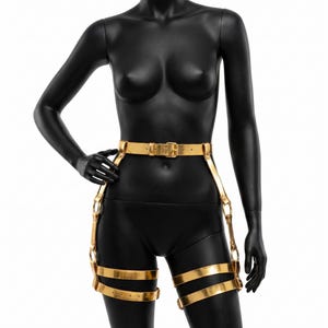 Puede incluir: Arn&eacute;s corporal dorado con cintur&oacute;n y correas para las piernas. El arn&eacute;s se muestra en un maniqu&iacute; negro. El cintur&oacute;n tiene una hebilla rectangular y las correas de las piernas est&aacute;n conectadas con anillos met&aacute;licos.
