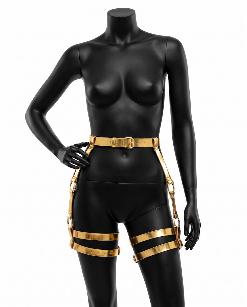 Puede incluir: Arn&eacute;s corporal dorado con cintur&oacute;n y correas para las piernas. El arn&eacute;s se muestra en un maniqu&iacute; negro. El cintur&oacute;n tiene una hebilla rectangular y las correas de las piernas est&aacute;n conectadas con anillos met&aacute;licos.