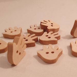 May include: A selection of light brown wooden hand-shaped buttons. Each button features two small holes for sewing. The buttons are scattered on a light-coloured surface, with varying shadows.