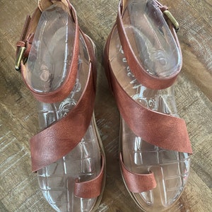 May include: A pair of brown leather sandals with ankle straps and a toe ring. The sandals have a clear plastic insole and a gold buckle.