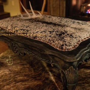 May include: A black coffee table with ornate carvings and a cowhide top. The table has a distressed finish and is accented with brown nailhead trim. The table is sitting on a cowhide rug.