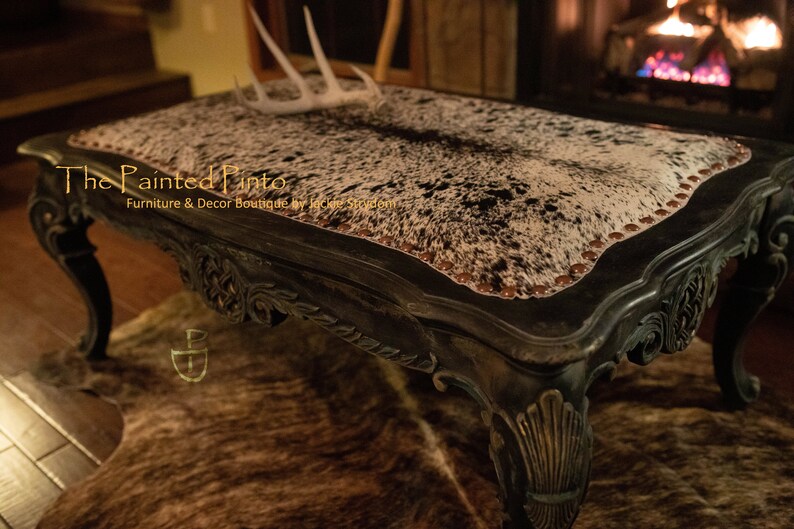 May include: A black coffee table with ornate carvings and a cowhide top. The table has a distressed finish and is accented with brown nailhead trim. The table is sitting on a cowhide rug.
