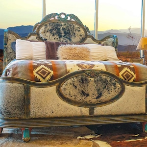 May include: A large, ornate bed frame with a cowhide headboard and footboard. The bed is covered in a colorful blanket with a geometric pattern. The bed is set against a backdrop of a desert landscape.