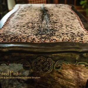 May include: A dark green, ornate table with a cowhide top. The table has a decorative, carved design on the legs and a row of copper-coloured studs around the edge of the top. The table is labelled "The Painted Pinto, Furniture & Decor Boutique by Jackie Strydom".