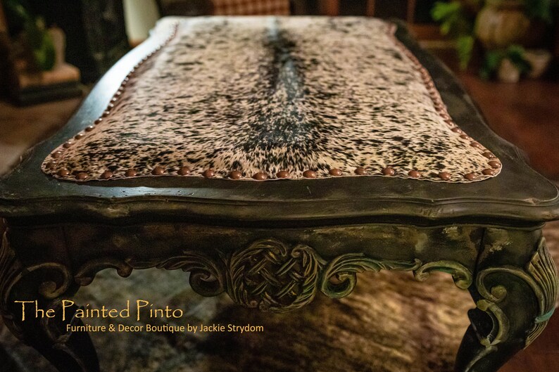 May include: A dark green, ornate table with a cowhide top. The table has a decorative, carved design on the legs and a row of copper-coloured studs around the edge of the top. The table is labelled "The Painted Pinto, Furniture & Decor Boutique by Jackie Strydom".