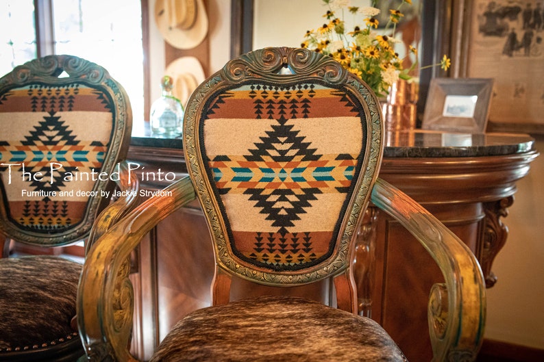 May include: Two ornate wooden chairs with a Southwestern-style patterned fabric upholstery. The chairs have a distressed paint finish and are decorated with gold accents. The chairs are set against a backdrop of a wooden bar with a granite countertop.