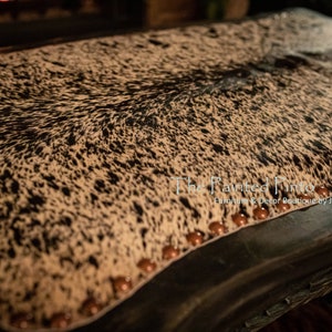 May include: A black and white cowhide ottoman with brown nailhead trim. The ottoman is upholstered in a black and white cowhide pattern. The ottoman is sitting on a dark wood floor.