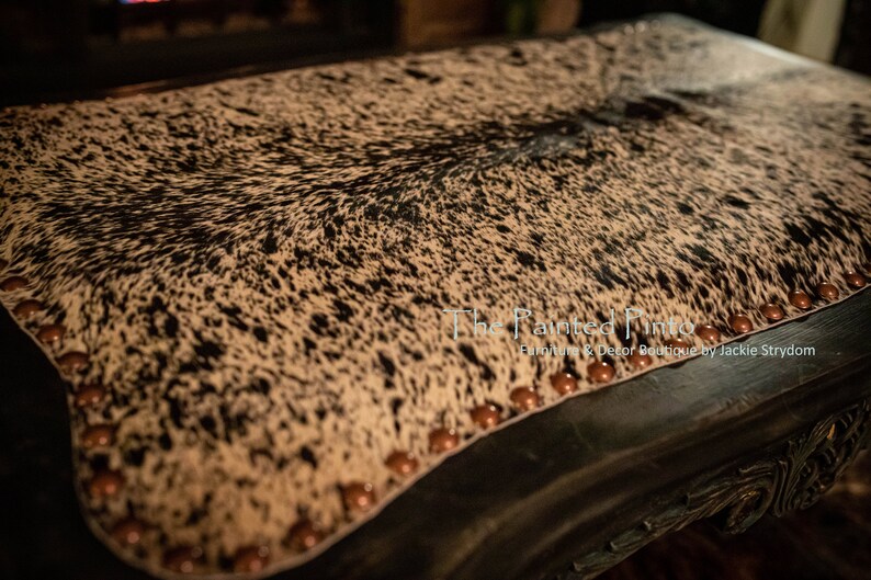 May include: A black and white cowhide ottoman with brown nailhead trim. The ottoman is upholstered in a black and white cowhide pattern. The ottoman is sitting on a dark wood floor.