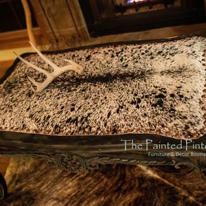 May include: A black coffee table with a cowhide top and decorative trim. The table has a distressed finish and features a white antler on top. The table is labelled "The Painted Pinto" with the text "Furniture & Decor Boutique by Jackie Strydom".