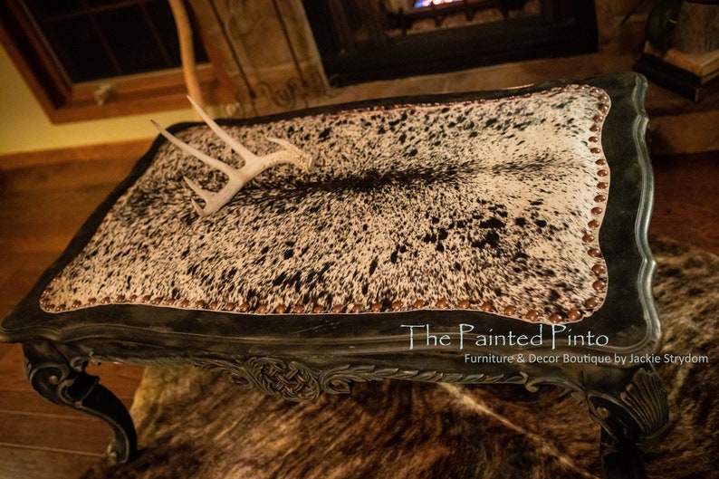 May include: A black coffee table with a cowhide top and decorative trim. The table has a distressed finish and features a white antler on top. The table is labelled "The Painted Pinto" with the text "Furniture & Decor Boutique by Jackie Strydom".