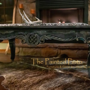 May include: A dark green coffee table with ornate carvings and gold accents. The table is sitting on a brown and white cowhide rug. The text "The Painted Pinto Furniture & Decor Boutique by Jackie Strydom" is visible in the bottom right corner of the image.