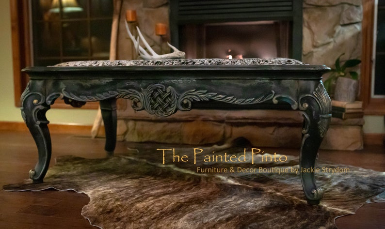 May include: A dark green coffee table with ornate carvings and gold accents. The table is sitting on a brown and white cowhide rug. The text "The Painted Pinto Furniture & Decor Boutique by Jackie Strydom" is visible in the bottom right corner of the image.