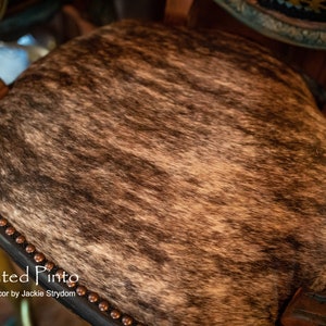 May include: Close-up of a brown and white cowhide seat with a black leather border and copper-colored studs. The seat is on a wooden chair.