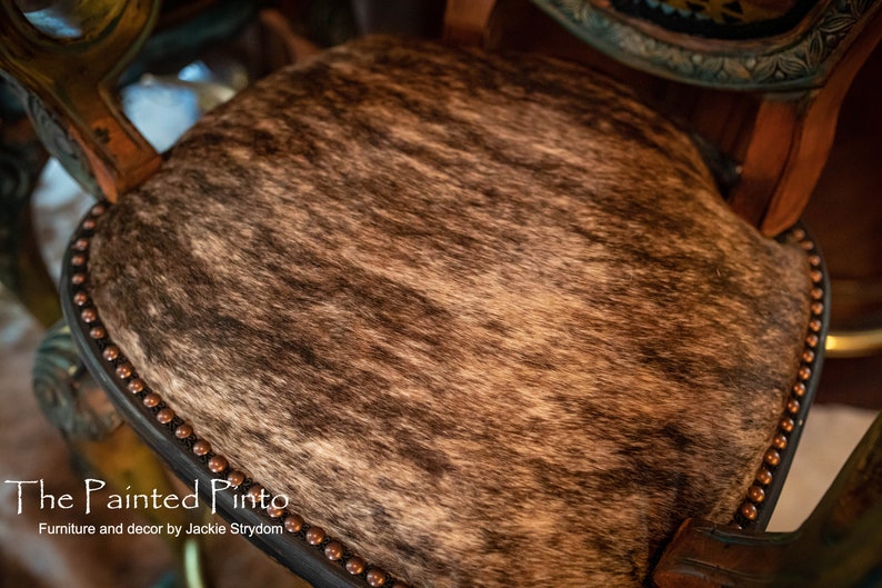May include: Close-up of a brown and white cowhide seat with a black leather border and copper-colored studs. The seat is on a wooden chair.