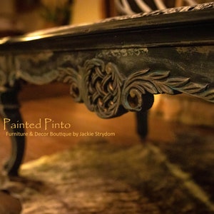 May include: A dark wood coffee table with intricate carvings and a distressed finish. The table has a carved design on the bottom edge and a single leg is visible in the foreground. The text "The Painted Pinto" is visible in the lower left corner of the image.