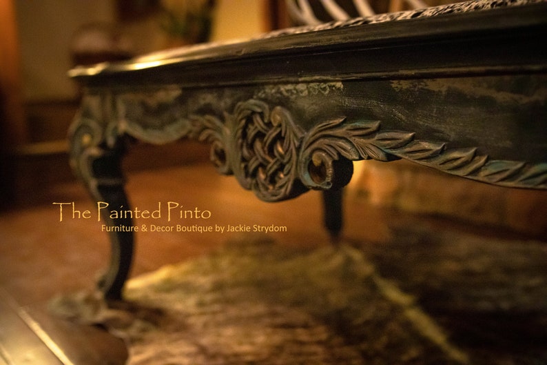 May include: A dark wood coffee table with intricate carvings and a distressed finish. The table has a carved design on the bottom edge and a single leg is visible in the foreground. The text "The Painted Pinto" is visible in the lower left corner of the image.