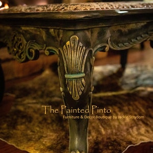 May include: A close-up of a dark wood coffee table with a decorative carved leg. The table has a distressed finish with gold accents. The table is sitting on a brown and white fur rug. The text "The Painted Pinto Furniture & Decor Boutique by Jackie Strydom" is visible below the table.