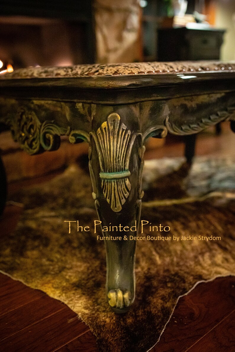 May include: A close-up of a dark wood coffee table with a decorative carved leg. The table has a distressed finish with gold accents. The table is sitting on a brown and white fur rug. The text "The Painted Pinto Furniture & Decor Boutique by Jackie Strydom" is visible below the table.