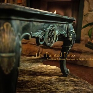May include: A dark green coffee table with ornate carvings and gold accents. The table is sitting on a cowhide rug. The text "The Painted Pinto" is visible in the image.