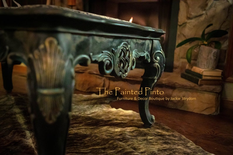 May include: A dark green coffee table with ornate carvings and gold accents. The table is sitting on a cowhide rug. The text "The Painted Pinto" is visible in the image.