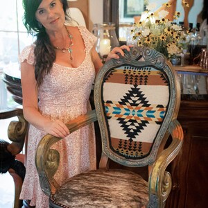 May include: A woman stands next to a wooden chair with a cowhide seat and a colorful patterned back. The chair is decorated with gold paint and has a carved design. The chair is labeled "The Painted Pinto" and "Furniture and decor by Jackie Strydom".