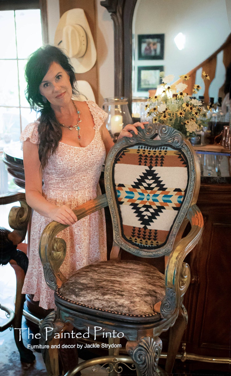 May include: A woman stands next to a wooden chair with a cowhide seat and a colorful patterned back. The chair is decorated with gold paint and has a carved design. The chair is labeled "The Painted Pinto" and "Furniture and decor by Jackie Strydom".