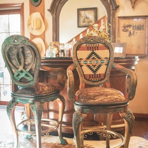 May include: Two ornate bar stools with cowhide upholstery and intricate carvings. The stools are painted in a distressed green and gold finish. The stool on the right has a colorful southwestern patterned fabric on the back.
