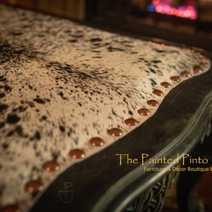 May include: Close-up of a black and white cowhide table top with copper nailhead trim. The table has a decorative carved edge and the text "The Painted Pinto Furniture & Decor Boutique by Jackie Strydom" is visible in the lower right corner.
