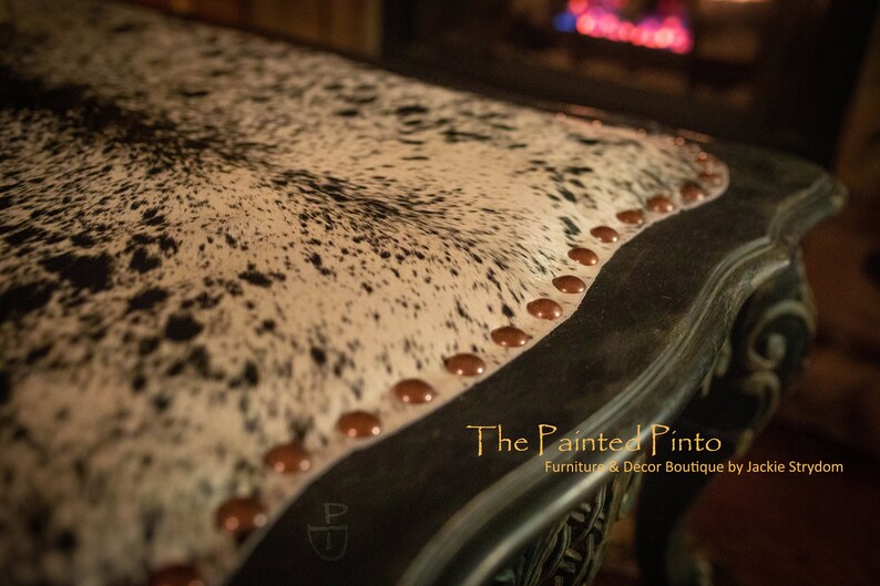 May include: Close-up of a black and white cowhide table top with copper nailhead trim. The table has a decorative carved edge and the text "The Painted Pinto Furniture & Decor Boutique by Jackie Strydom" is visible in the lower right corner.