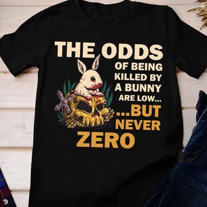 Puede incluir: Camiseta negra con un gráfico de una calavera y un conejito con el texto "The Odds of Being Killed by a Bunny Are Low... But Never Zero."
