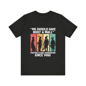Peut inclure: T-shirt noir avec un motif graphique repr&eacute;sentant quatre silhouettes am&eacute;rindiennes devant des rectangles color&eacute;s. Le texte se lit "WE SHOULD HAVE BUILT A WALL" et "FIGHTING ILLEGAL IMMIGRATION SINCE 1492."
