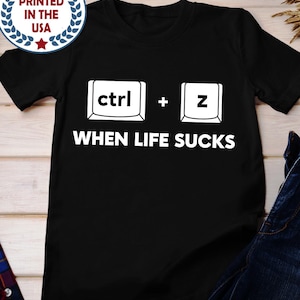 Puede incluir: Camiseta negra con el texto "WHEN LIFE SUCKS" debajo de un gráfico de las teclas "ctrl" y "z". La camiseta también tiene un logotipo de "PRINTED IN THE USA". La camiseta está hecha de un material suave.