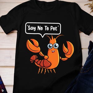 May include: Black t-shirt with a cartoon lobster in orange, with large claws and googly eyes. The lobster has a speech bubble with the text "Say No To Pot". The shirt is a casual style.